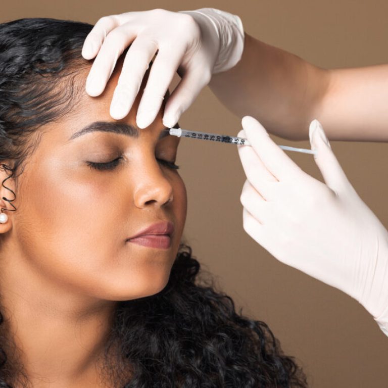 Cosmetologist making botox injection in interbrow zone to latin chubby woman getting anti-wrinke treatment over brown studio background, closeup